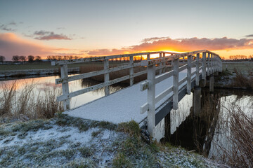 White wooden arched bridge with a layer of snow in the morning sun. Behind Lake Dirkshorn lies this...