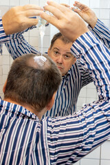 A man in pajamas applies a preparation for baldness in the bathroom in front of the mirror