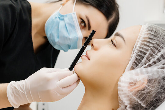 Beauticians in white gloves draw on the face with a pencil, preparing to inject a rejuvenating facial filler into a young woman. Injection beauty procedure.