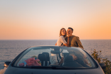 Beautiful happy beloved couple posing to a black convertible on the shore of the Mediterranean sea...