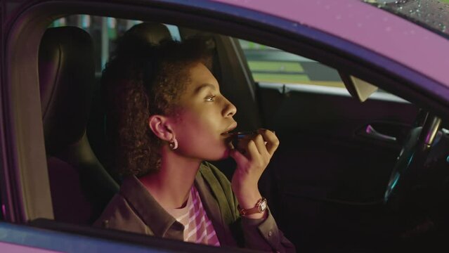 Side Through Window Footage Of Young Black Woman Sitting On Passenger Seat In Car And Putting On Lip Balm While Waiting For Driver At Night