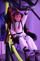 Little adorable girl with long hair European appearance sits on virtual reality attraction Cosmos in white cosmonaut costume closes up attraction with hand