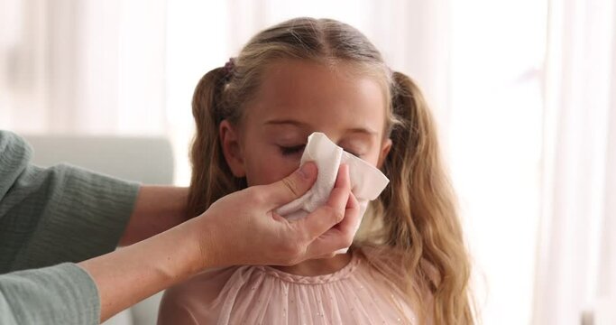 Mother helps preschooler girl blow snot into napkin. Illness and treatment at home. Care and support from parent to child in bright room at home