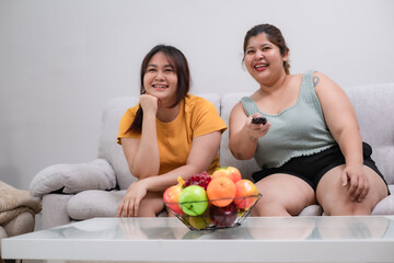 Obraz na płótnie Canvas Two young overweight women sitting on sofa in living room looking television with smile and happiness emotion at home.