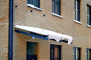 Fototapeta na wymiar Glass canopy covered with snow after snow storm. Door canopy with support construction, glass panel mounted with tension rod and wall brackets on building facade. Snow and icicles on roof