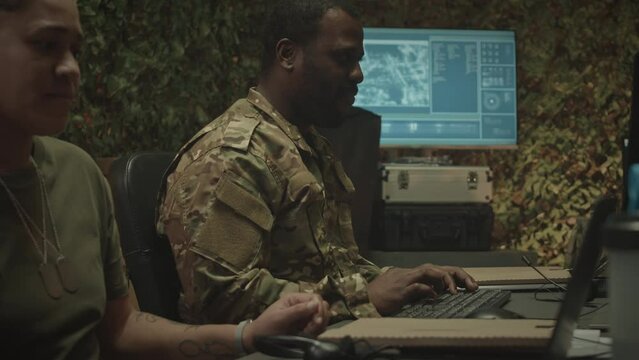 Tilt up shot of two diverse colleagues in camouflage uniform sitting at desk in front of computer and laptop, typing and observing monitoring programs while working in military control center at night