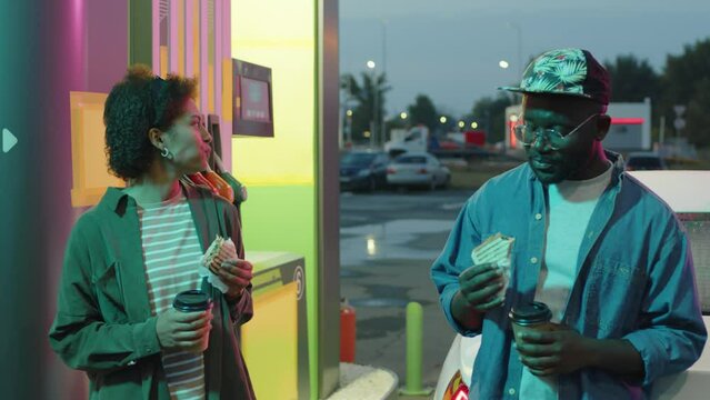 Medium footage of African American cross-sex friends having sandwich and coffee and speaking during rest stop at petrol station at night