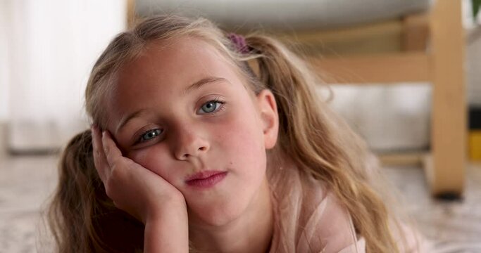Tired girl with blonde hair lies on floor with cheek resting on hand in living room. Boredom and fatigue in country house. Child looks around slow motion
