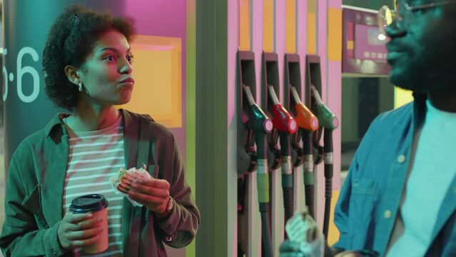 Medium footage of Black young lady standing near gasoline pump and having snack while speaking to male friend or boyfriend at neon lit gas filling station at night