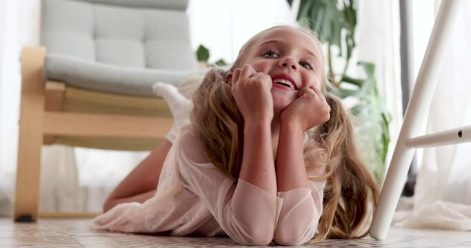 Smiling girl lies on floor with cheeks resting on hands against armchair in light living room. Cheerful child looks around and smiles slow motion