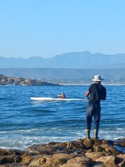 Fototapeta na wymiar Plettenberg Bay, Eastern Cape Local fisherman fishing in the sea, whilst standing on rocks and a person in a canoe rowing past. Mountains in the background