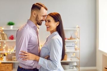 Portrait of a young laughing smiling man and woman hugging each other indoors standing in the living room at home. Happy couple in love. Relationships, romance and Valentines day concept.