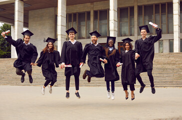 Happy smiling graduates students classmates jumping in black graduate gown with diploma in hands and celebrating graduation in university campus outdoors. Portrait of graduated guys and girls.