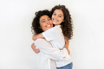 Happy young mother hugging her daughter and smiling at camera, expressing tenderness, love and care, standing over white studio wall background