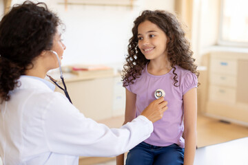 Fototapeta premium Female pediatrician examining child girl patient with stethoscope, doing pediatric checkup in hospital children medical care concept