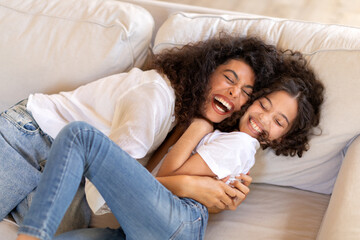 Mom tickling her daughter and laughing while having fun together on sofa, mother and child girl...