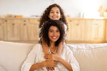 Portrait of cute daughter embracing her mother from the back, sitting on sofa, smiling together to...
