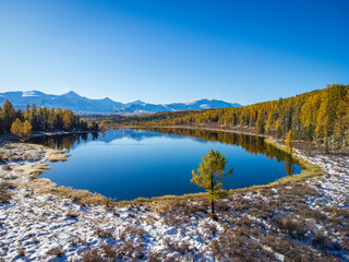 Vivid aerial shot of a serene mountain lake surrounded by autumn trees with snowy patches in Altai...
