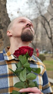 Handsome bearded and fashionable handsome man holding a bouquet of flowers and dreaming about his desires looking up outdoors. Dreamy seducer of women, sexy man with roses. Vertical