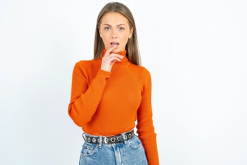 Nervous puzzled young beautiful girl wearing orange turtleneck opens mouth from surprise, reacts on sudden news.