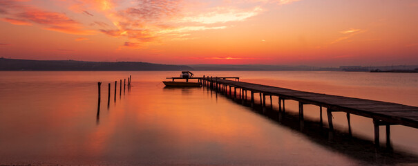 Fototapeta premium Scenic view of exciting red sunset at the shore with a wooden pier and moored boat
