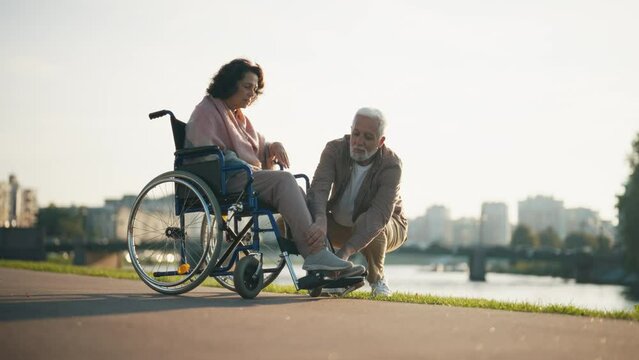Husband walking with wife in wheelchair. Senior family couple spending time in city park near pond. Man helped woman showing love care, they smiling. Living with physical disability concept.
