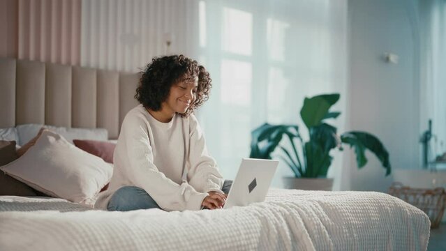 Contented african american girl reading typing messages on laptop chatting with friends sitting in bed at home. Smiling positive curly relaxed female enjoying online communication on computer.
