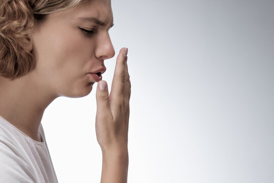Bad breath. Young girl checking her breath with her hand, blowing to it, standing over grey background. Bad smell from the mouth, toothache, having problems with teeth