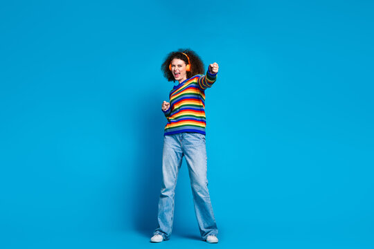 Full size photo of attractive young woman music earphones fight fist wear trendy rainbow print clothes isolated on blue color background