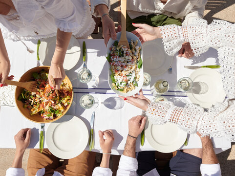 An overhead shot of hands serving vibrant salads at a sunny outdoor table, with glasses of white wine awaiting the start of a festive meal.