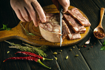 The cook cuts fresh brawn into pieces with a knife on a kitchen wooden board before preparing...