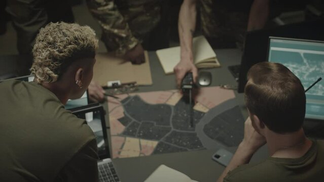 Top view shot of multiethnic military workers wearing camouflage uniform standing by table with satellite map on it and having discussion, working together in dark military surveillance office