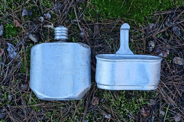 a set of an old white aluminum army flask and a kettle lie on the ground and green moss on the...