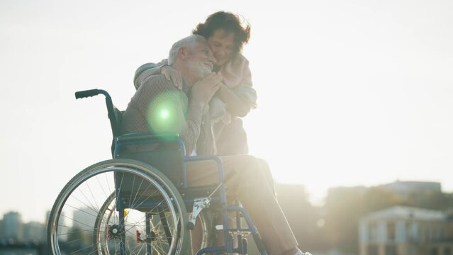 Wife walking with husband in wheelchair. Senior family couple spending time in city park near pond. Woman hugging man show love care, they happiness talking. Living with physical disability concept.