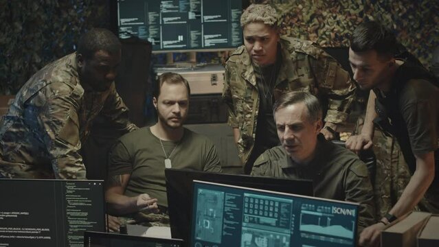 Medium shot of ethnically diverse team of male and female military cyber security workers looking at data on computer display and having discussion in dark office at night time
