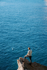 Fototapeta na wymiar Young man stands on a stone ledge above the sea and looks into the distance. Back view