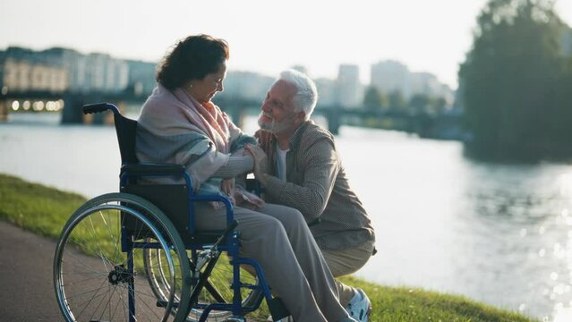 Husband walking with wife in wheelchair. Senior family couple spending time in city park near pond. Man hugging female showing love care, they smiling. Living with physical disability concept.