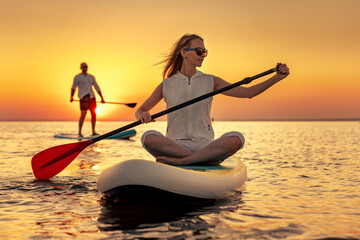 Young man and woman walks with sup boards at sunset lake. Beautiful girl is sitting on stand up paddle board at calm lake
