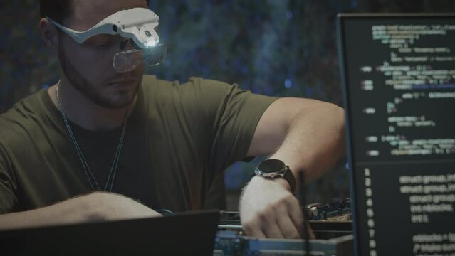 Waist up of Caucasian male military engineer in magnifying goggles with light assembling motherboard with programming code on computer display in focused foreground