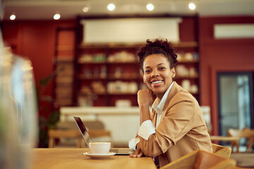 A smiling African woman, sitting at the cafe, working over the laptop.