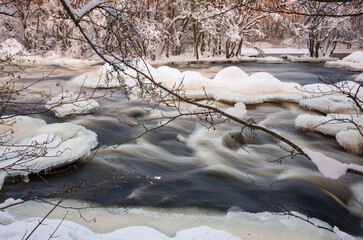 Fast river flows among icy stones in winter forest surrounded by trees covered with snow,...