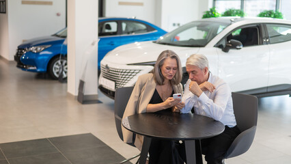 A mature couple is sitting in a car dealership and looking at a smartphone. 