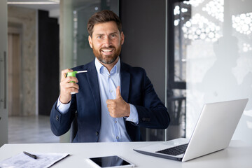 Portrait of a smiling young man sitting in a suit at a desk with a laptop, holding a cough spray...