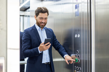 A young smiling businessman and lawyer is standing in the elevator of an office building and pressing the wrong button while using a mobile phone