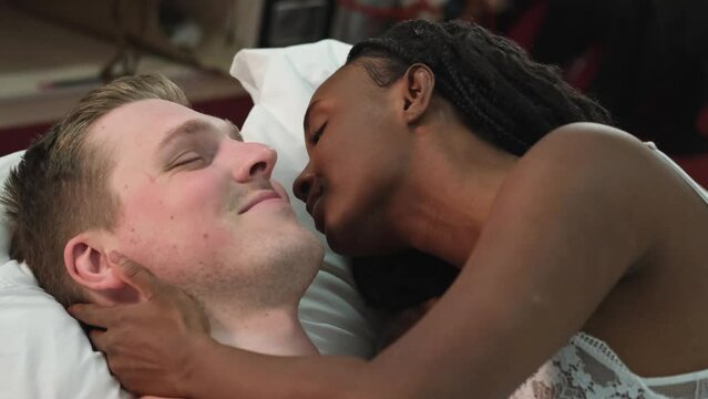 Sensual multiethnic couple kissing and hugging in bed at home. Romantic guy and girl having a good time together in the bedroom. A beautiful wife and a handsome husband look at each other, enjoy a