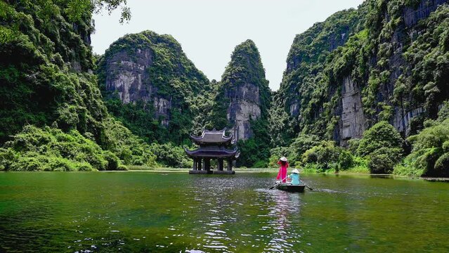 4k video,Asian women wearing red traditional Vietnamese cultural clothing on a boat floating in the river flowing through the mountains at Trang An, Vietnam.