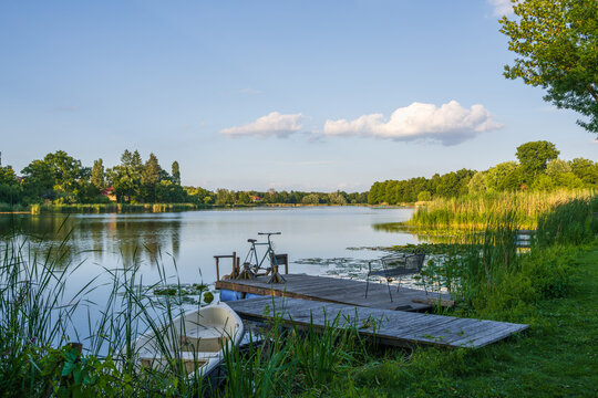 Country summer motif. Small lake, green shore. A wooden pier extending into the water with a homemade bicycle. White boat in the water. Sunny day. Falkensee near Berlin, Germany