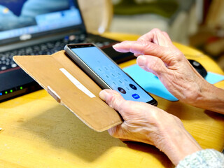 ノートパソコンの前でスマホの番号を操作する高齢女性の手元 - obrazy, fototapety, plakaty