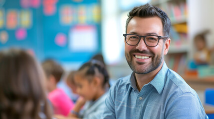 Obraz premium Happy male elementary school teacher. Students are in classroom behind him in a blurry background. Fun and enjoyable learning, love for education concept.