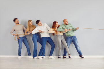 Group of happy friends playing tug of war and having fun together. Team of confident strong young adult people standing by grey color wall background, holding rope and pulling it in one direction - obrazy, fototapety, plakaty